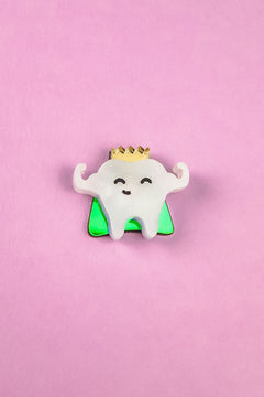 A cute Tooth King Pin enamel pin features a smiling tooth with a gold crown, arms raised in front of a green triangle on a pink background. This handcrafted accessory is perfect for adding royal flair to your collection.