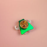 A small, green, triangular Tooth King Pin with a gold circular clasp and white accents sits on a soft pink surface—a charming handcrafted accessory.