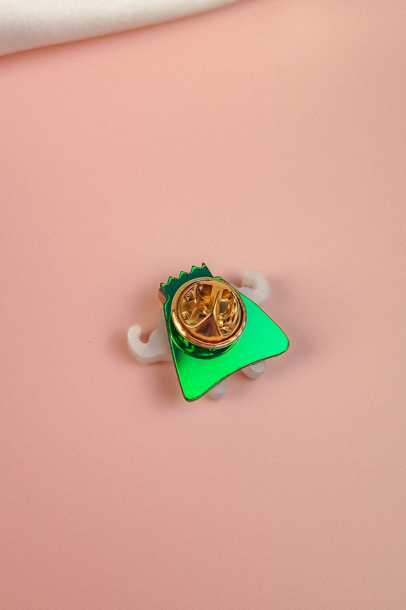 A small, green, triangular Tooth King Pin with a gold circular clasp and white accents sits on a soft pink surface—a charming handcrafted accessory.