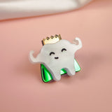 A cute handcrafted accessory, this enamel Tooth King Pin features a smiling tooth with little arms, a gold crown, and a shiny green base against a pink background with a white cloth in the corner.