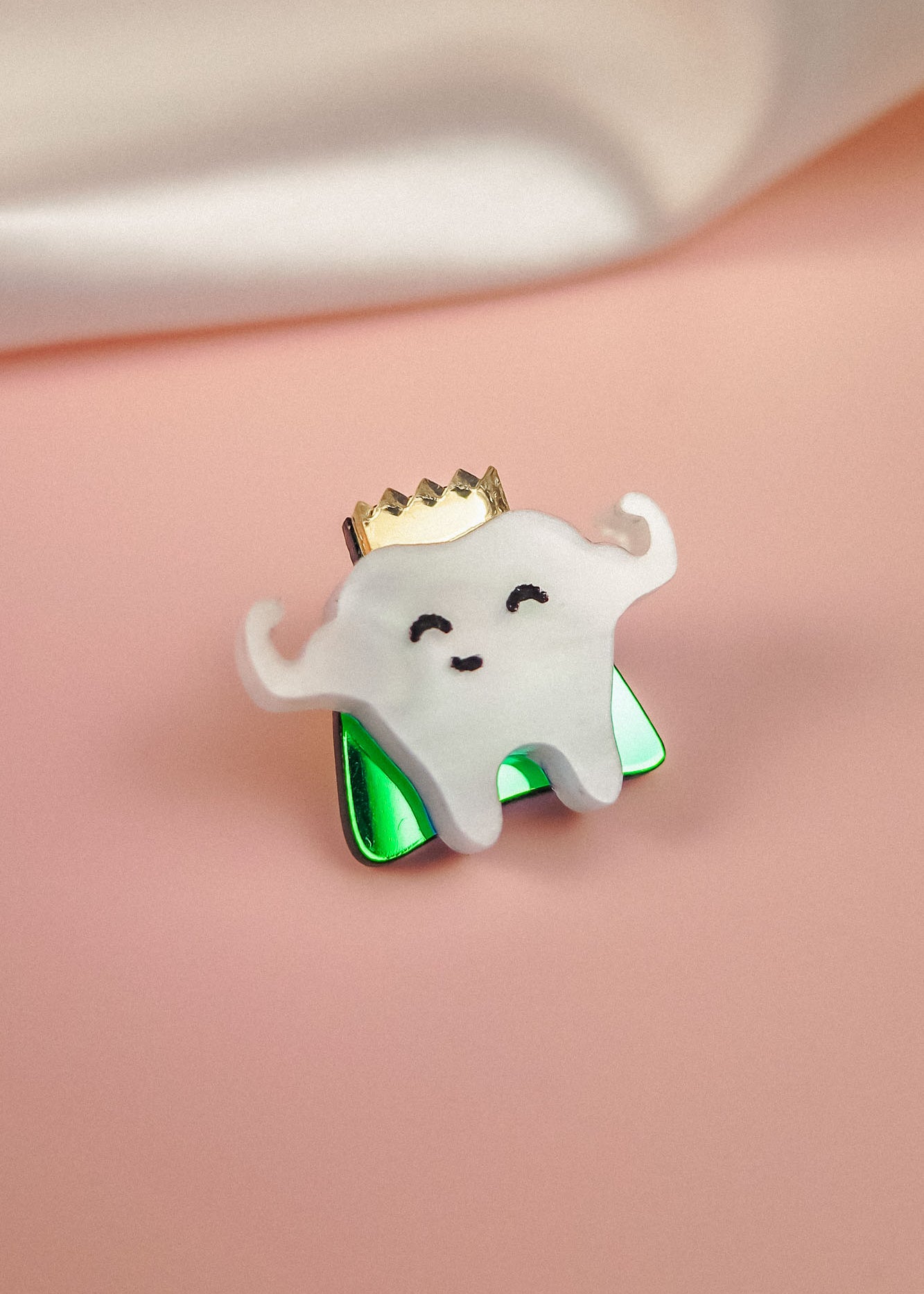 A cute handcrafted accessory, this enamel Tooth King Pin features a smiling tooth with little arms, a gold crown, and a shiny green base against a pink background with a white cloth in the corner.