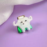 A cute Tooth King Pin shaped like a smiling tooth with a gold crown and flexed arms sits on a shiny green base, placed on a purple surface with white fabric in the background—a charming handcrafted accessory for any collection.
