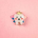 A smiling cartoon tooth with rosy cheeks, wings, and a gold crown sits centered on a pink background, making this handcrafted Tooth Fairy Pin perfect for celebrating childhood milestones.