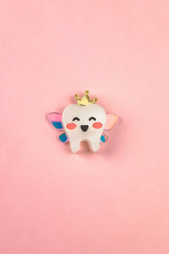 A smiling cartoon tooth with rosy cheeks, wings, and a gold crown sits centered on a pink background, making this handcrafted Tooth Fairy Pin perfect for celebrating childhood milestones.
