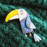 A handcrafted Toucan Brooch with a yellow beak and smiling face brings tropical charm as it rests on a textured dark green fabric background.