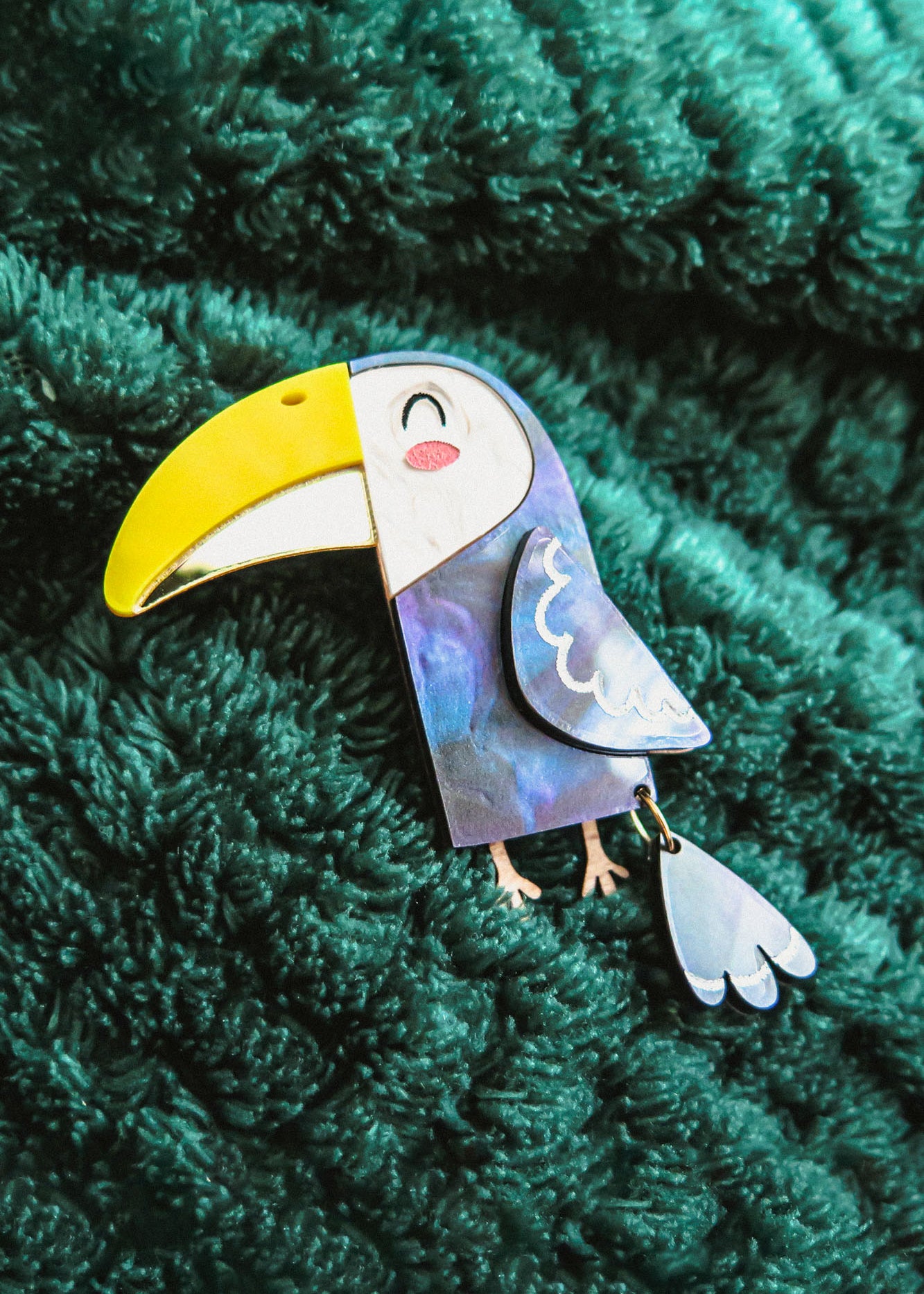 A handcrafted Toucan Brooch with a yellow beak and smiling face brings tropical charm as it rests on a textured dark green fabric background.