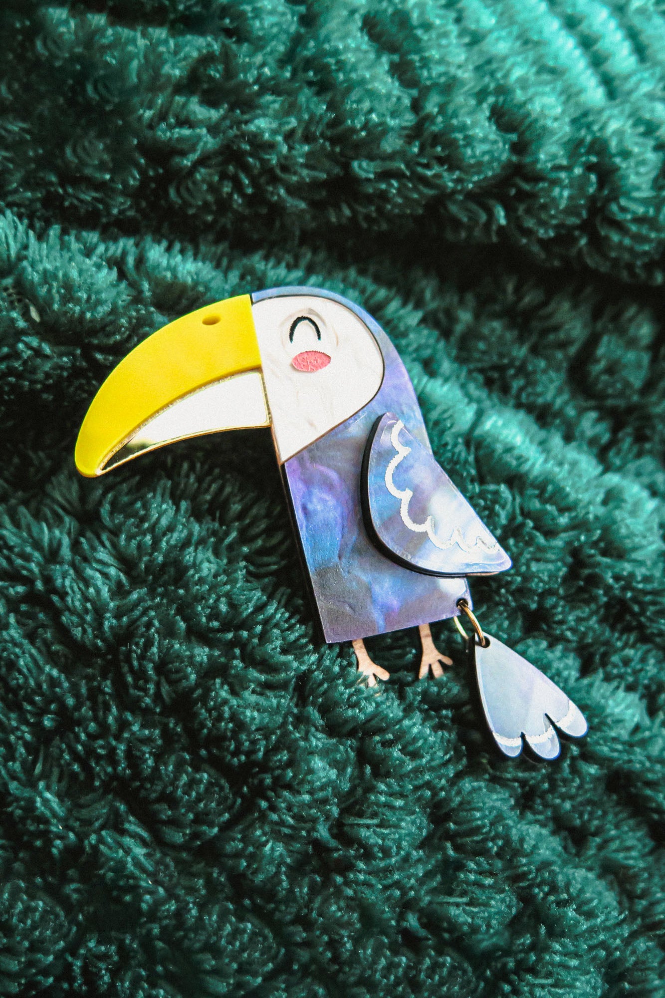 A handcrafted Toucan Brooch with a yellow beak and smiling face brings tropical charm as it rests on a textured dark green fabric background.