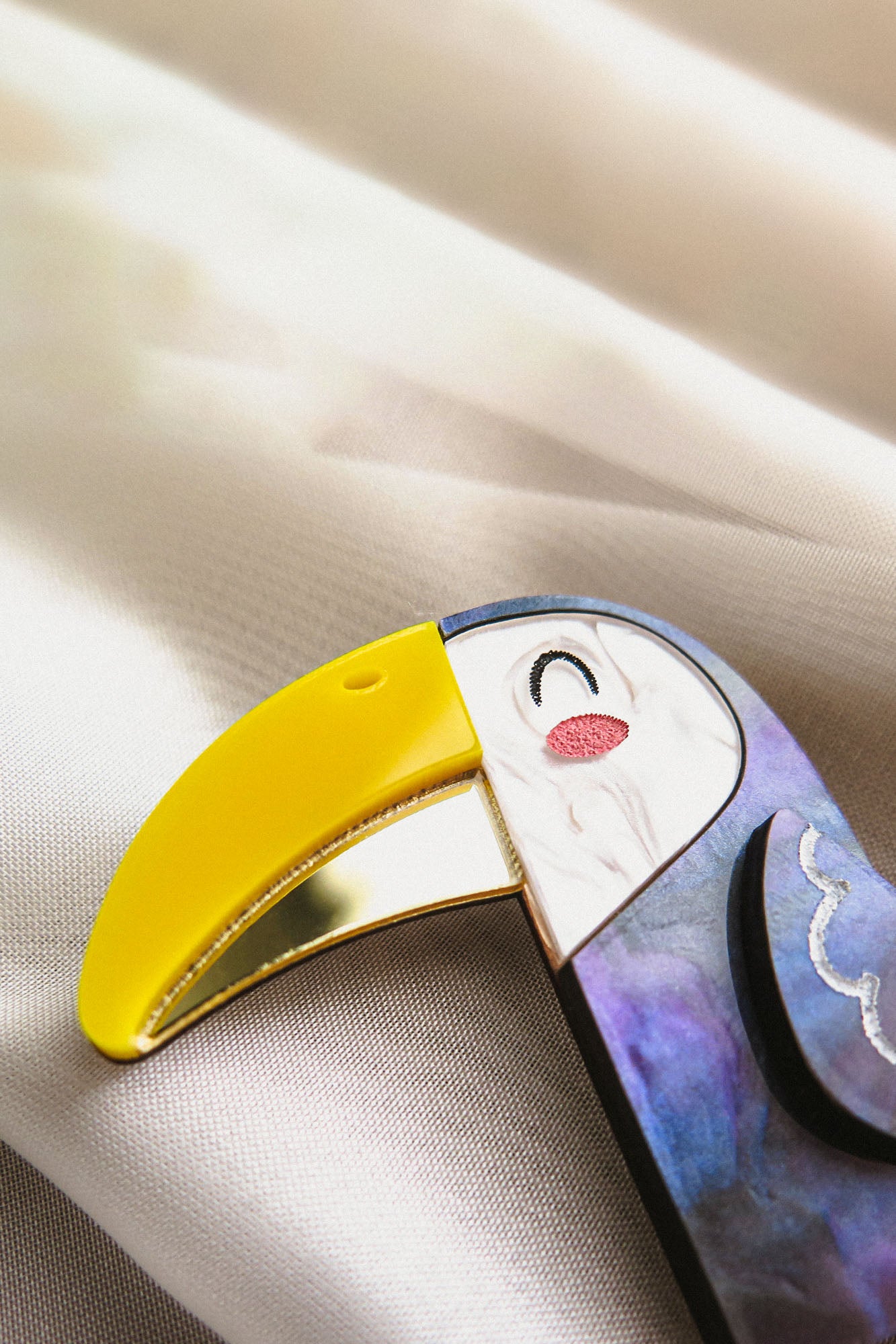 A close-up of a handcrafted Toucan Brooch with a yellow beak, white face, and vibrant painted details, resting on a soft, light-colored fabric background, radiating tropical charm.