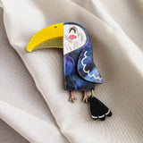 A handcrafted Toucan Brooch, featuring a bright yellow beak and a dangling tail, brings tropical charm as it rests on soft, cream-colored fabric.
