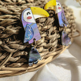 A pair of handcrafted Toucan Earrings with yellow beaks, purple wings, and dangling tail feathers adds a playful pop of color to a woven basket backdrop, surrounded by dried grasses and soft white fabric.