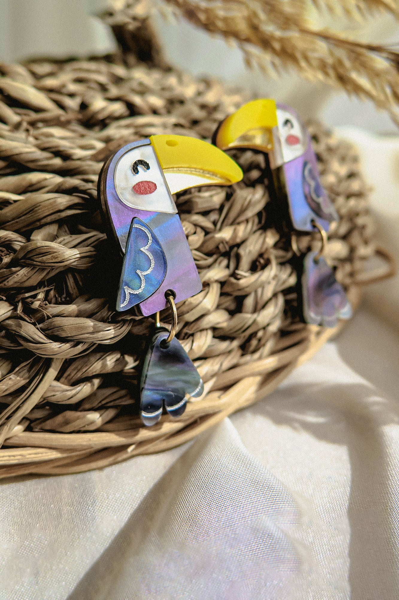 A pair of handcrafted Toucan Earrings with yellow beaks, purple wings, and dangling tail feathers adds a playful pop of color to a woven basket backdrop, surrounded by dried grasses and soft white fabric.