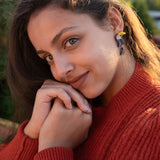 A young woman with long brown hair, wearing a red sweater and playful pop of color from handcrafted earrings, smiles softly while resting her chin on her hands outdoors in natural sunlight.