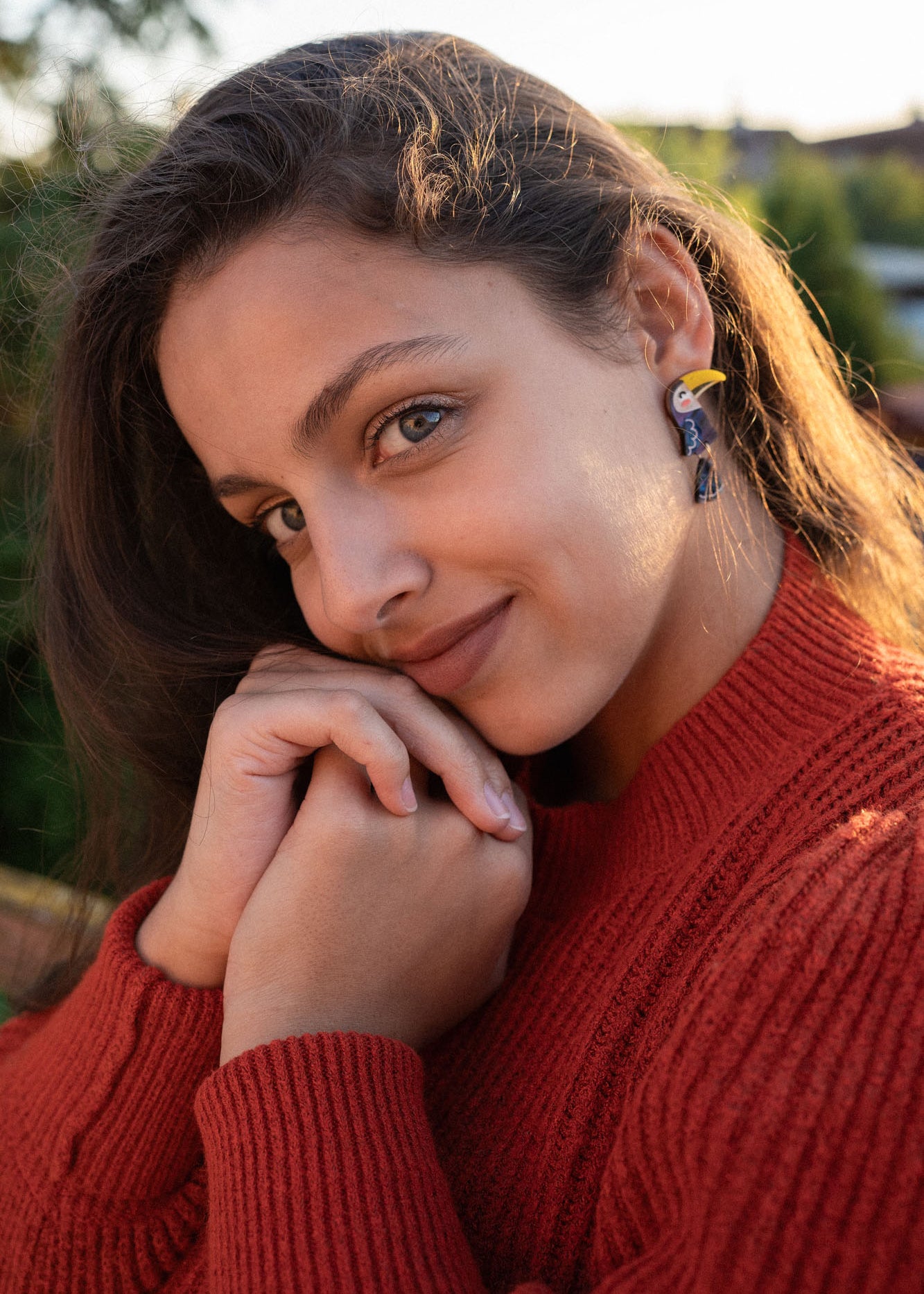 A young woman with long brown hair, wearing a red sweater and playful pop of color from handcrafted earrings, smiles softly while resting her chin on her hands outdoors in natural sunlight.
