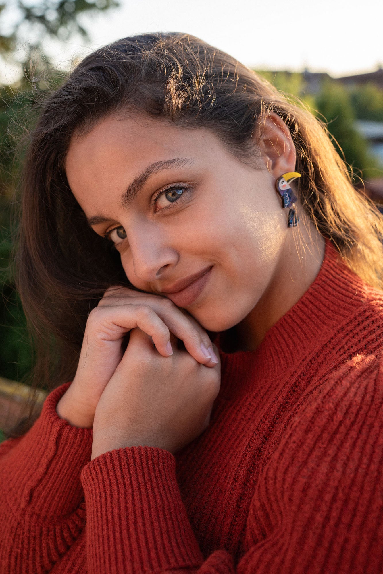 A young woman with long brown hair, wearing a red sweater and playful pop of color from handcrafted earrings, smiles softly while resting her chin on her hands outdoors in natural sunlight.