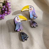 Two colorful Toucan Earrings made of iridescent materials rest on a light, satin-like fabric. Purple flowers are partially visible in the top left corner, adding to the playful pop of color in these handcrafted earrings.