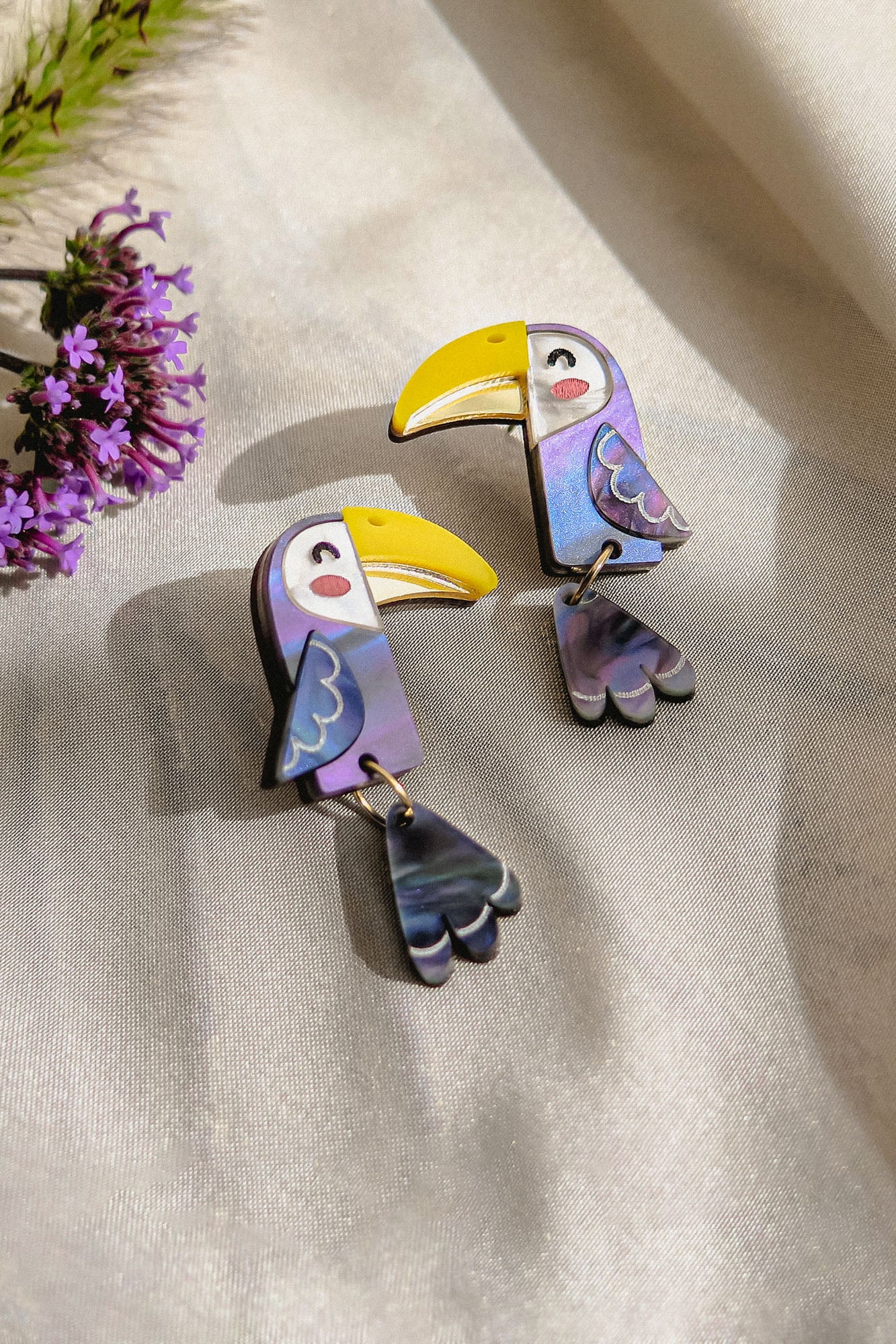Two colorful Toucan Earrings made of iridescent materials rest on a light, satin-like fabric. Purple flowers are partially visible in the top left corner, adding to the playful pop of color in these handcrafted earrings.