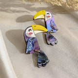 Handcrafted Toucan Earrings featuring yellow beaks, purple and blue wings, and dangling tails add a playful pop of color. Displayed on a light fabric background with dried pampas grass in the corner.