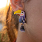 Close-up of a person’s ear adorned with handcrafted Toucan Earrings, featuring a whimsical yellow beak and dangling wing—a playful pop of color against the softly lit outdoor background.