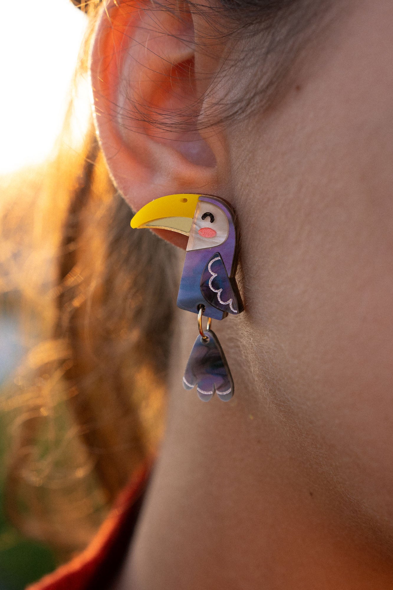 Close-up of a person’s ear adorned with handcrafted Toucan Earrings, featuring a whimsical yellow beak and dangling wing—a playful pop of color against the softly lit outdoor background.