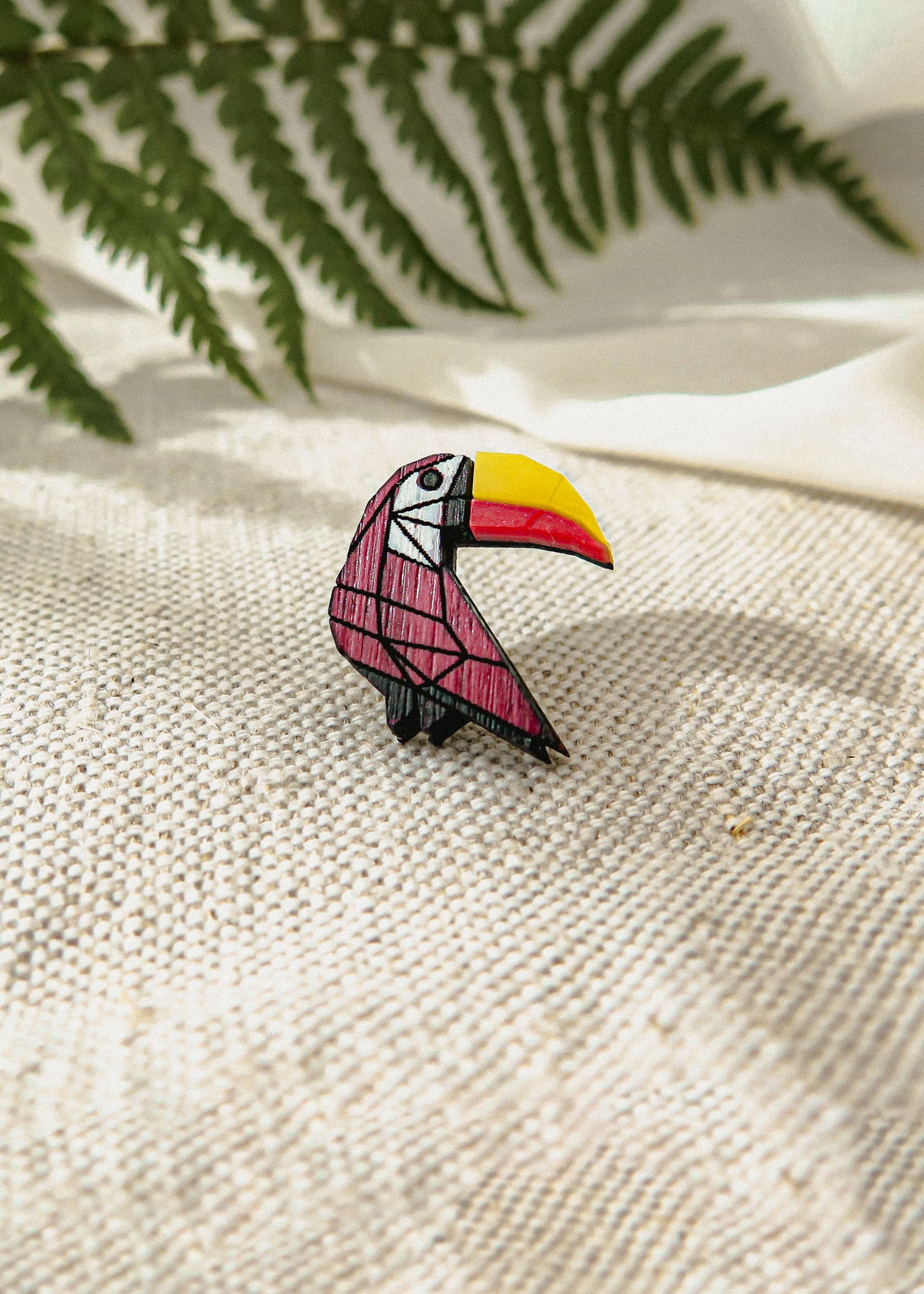 A handcrafted, colorful toucan pin with a yellow beak and red and black body rests on textured beige fabric, surrounded by green fern leaves and soft white cloth in the background.