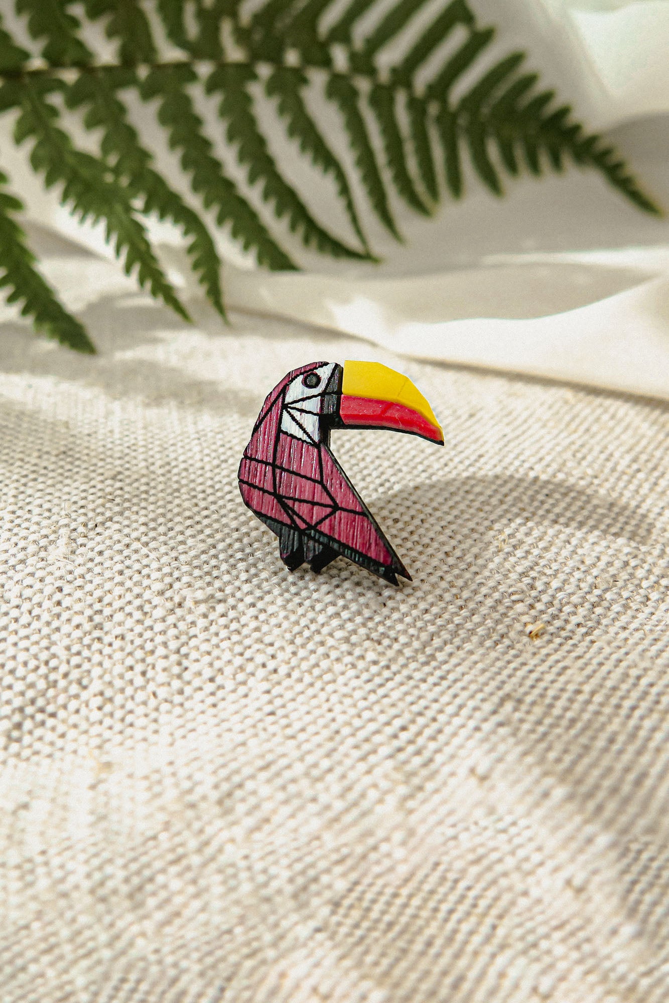 A handcrafted, colorful toucan pin with a yellow beak and red and black body rests on textured beige fabric, surrounded by green fern leaves and soft white cloth in the background.