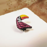 A colorful Toucan Pin brooch with a geometric, origami-inspired design rests on a white surface, featuring a vibrant yellow and orange beak and a pink body, handcrafted from eco-friendly materials.