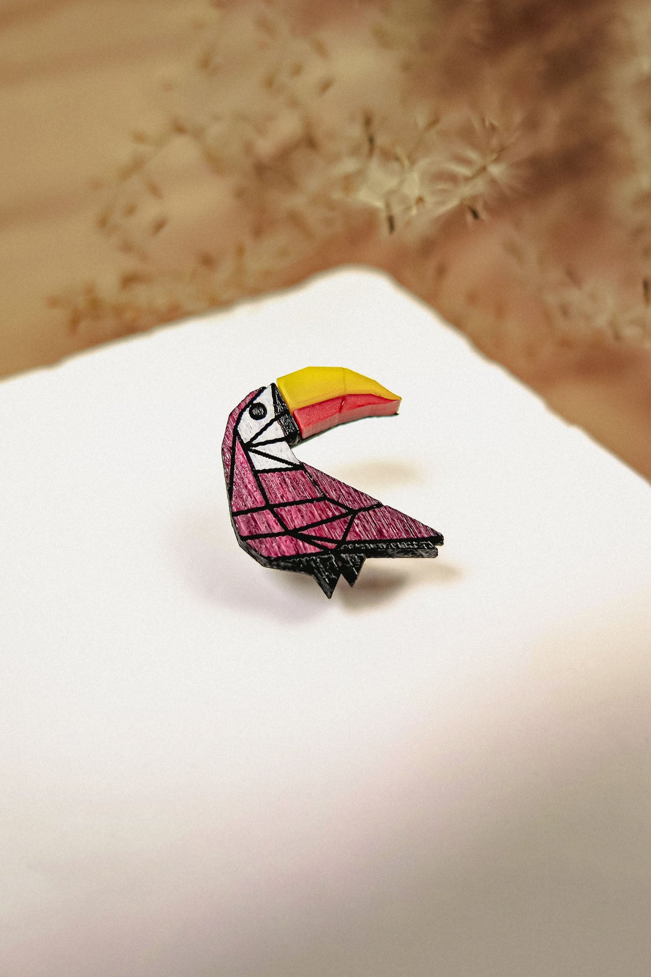A colorful Toucan Pin brooch with a geometric, origami-inspired design rests on a white surface, featuring a vibrant yellow and orange beak and a pink body, handcrafted from eco-friendly materials.