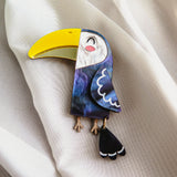 A handcrafted Toucan Brooch with a bright yellow beak and dark blue body captures tropical charm as it rests on soft, white, textured fabric.