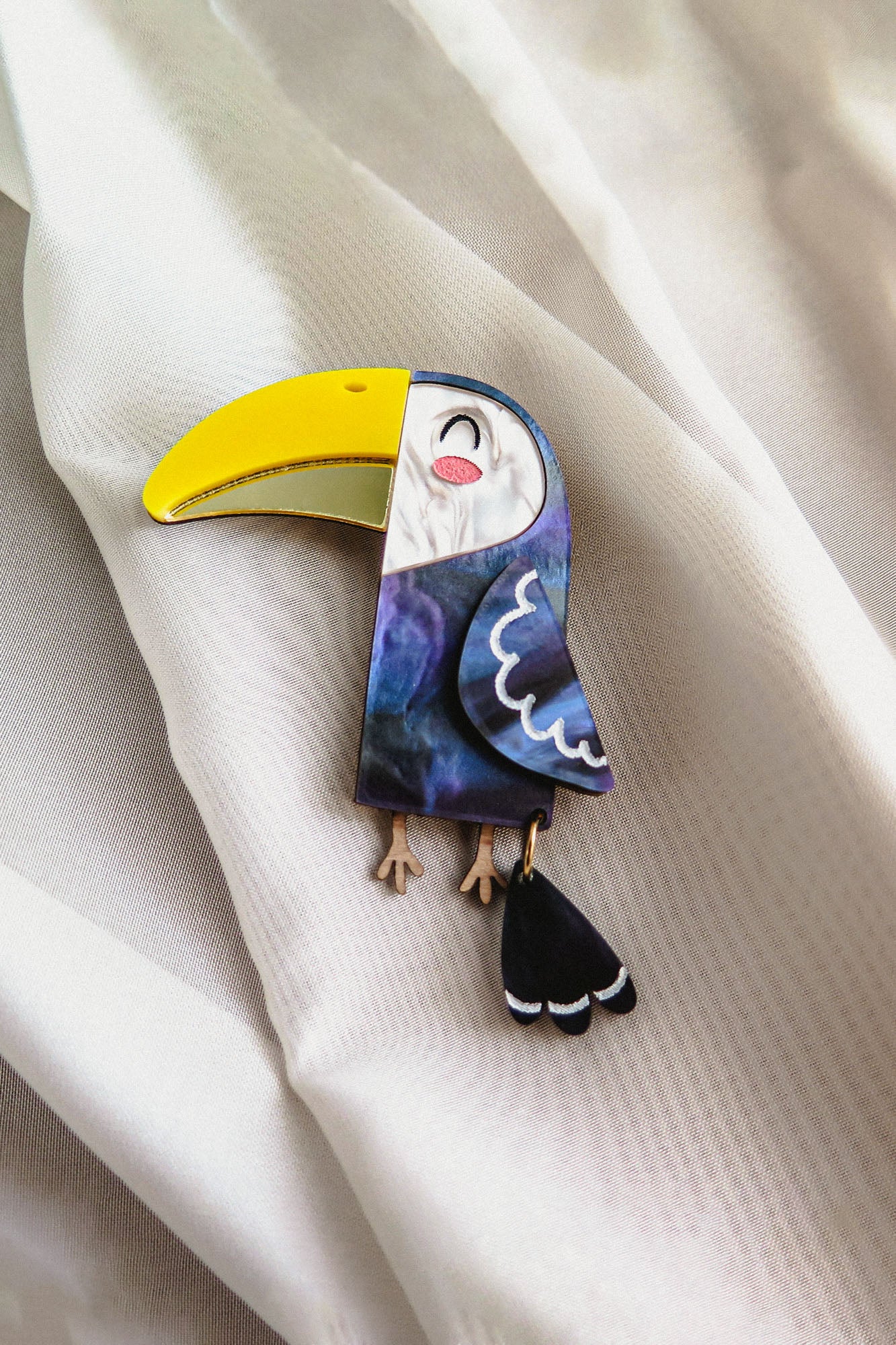A handcrafted Toucan Brooch with a bright yellow beak and dark blue body captures tropical charm as it rests on soft, white, textured fabric.