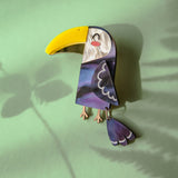 A handcrafted Toucan Brooch with a yellow beak and purple body lies on a green surface, radiating tropical charm as it casts a shadow alongside a fern leaf shadow.