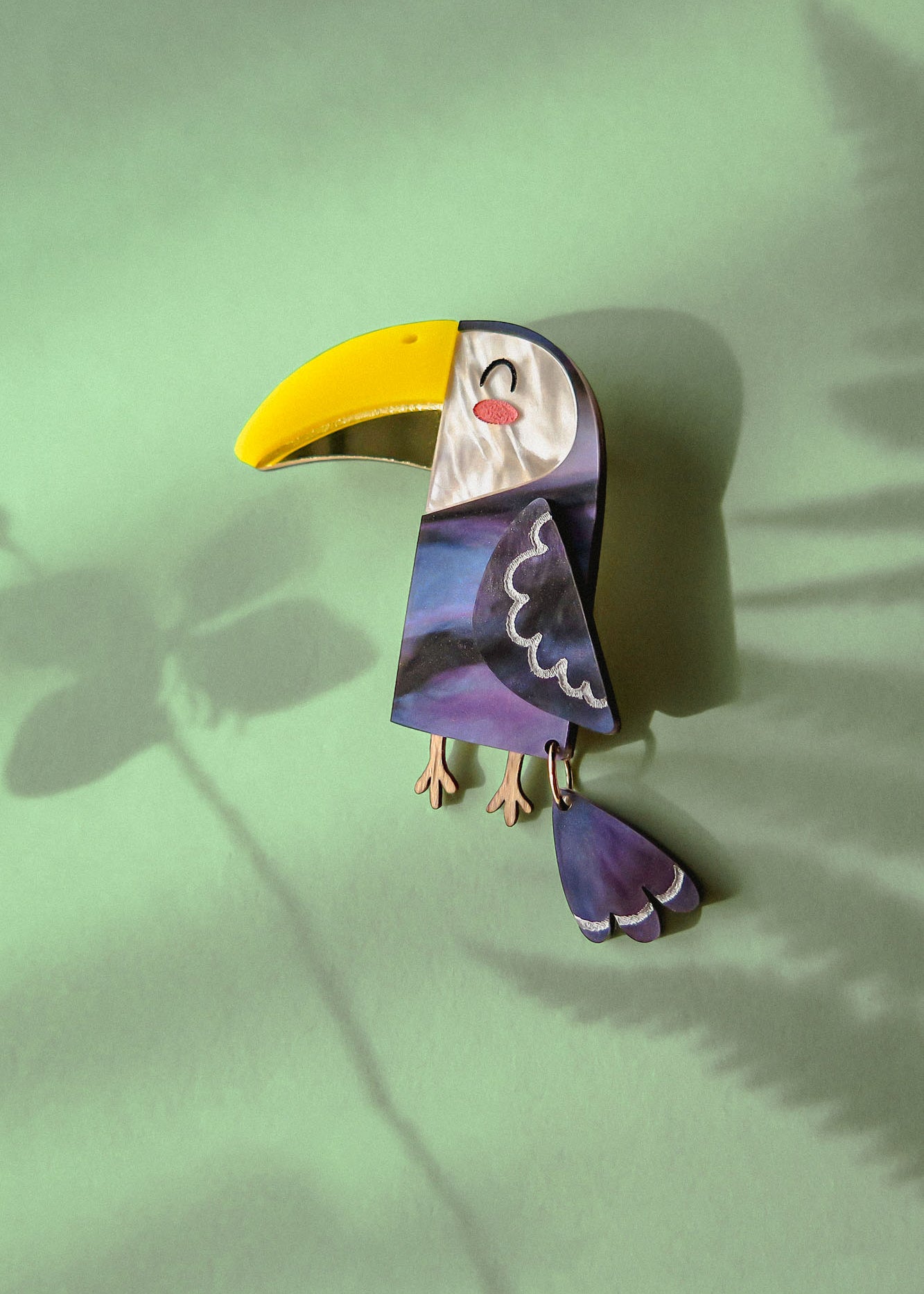 A handcrafted Toucan Brooch with a yellow beak and purple body lies on a green surface, radiating tropical charm as it casts a shadow alongside a fern leaf shadow.