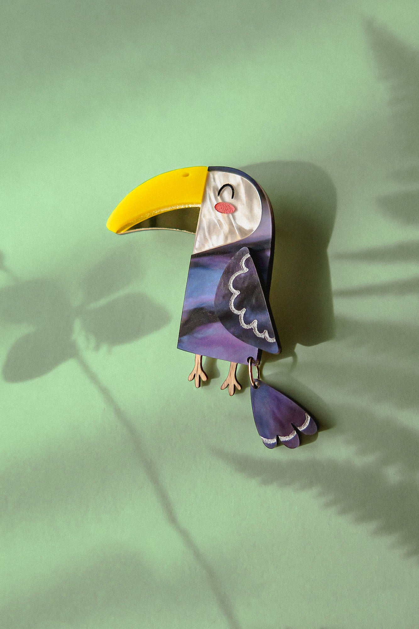 A handcrafted Toucan Brooch with a yellow beak and purple body lies on a green surface, radiating tropical charm as it casts a shadow alongside a fern leaf shadow.
