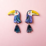 Add a playful pop of color to your look with these handcrafted Toucan Earrings, featuring yellow beaks, blue wings, and dangling blue tail feathers, displayed on a soft pink background.