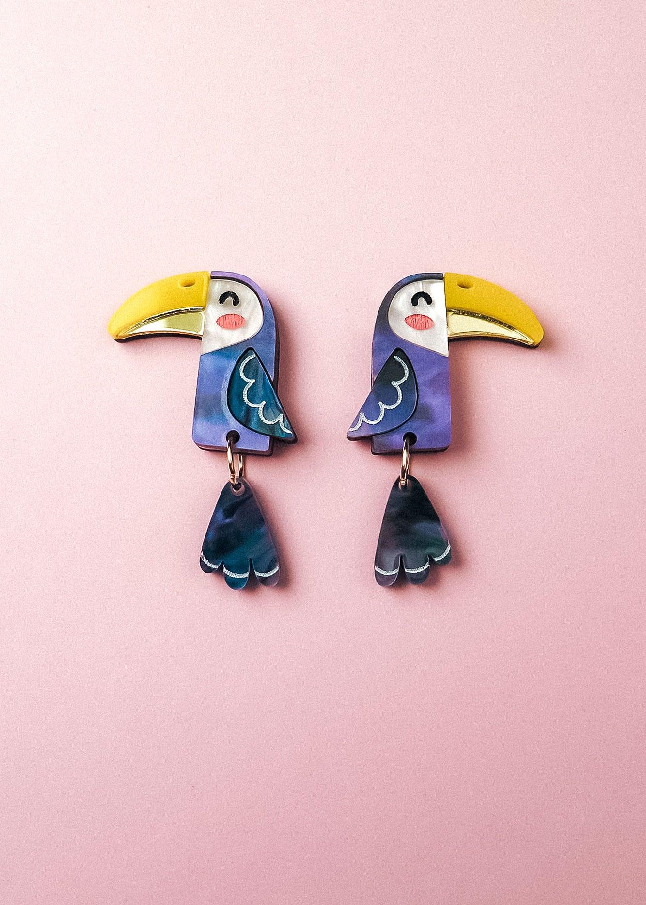 Add a playful pop of color to your look with these handcrafted Toucan Earrings, featuring yellow beaks, blue wings, and dangling blue tail feathers, displayed on a soft pink background.