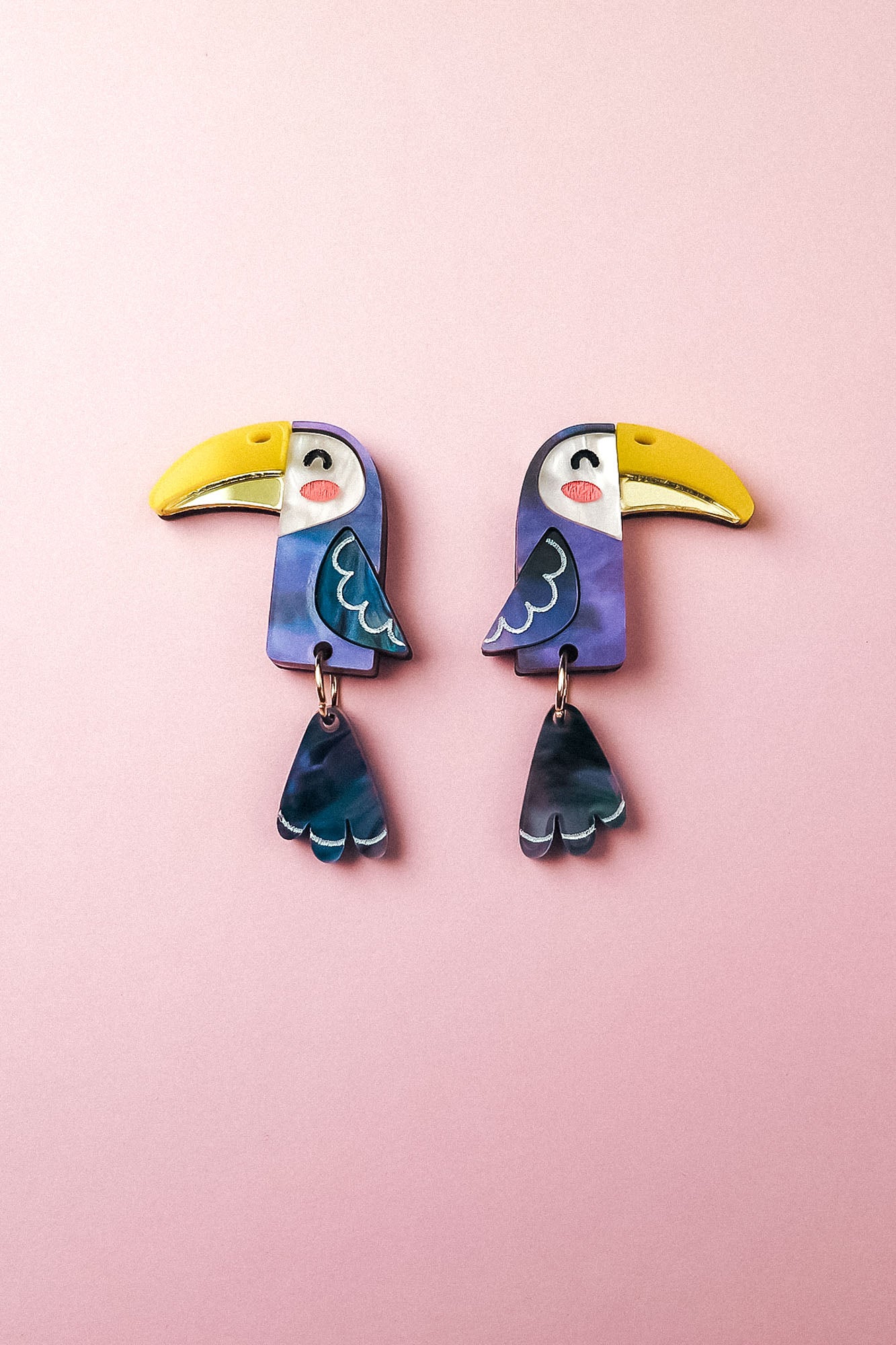 Add a playful pop of color to your look with these handcrafted Toucan Earrings, featuring yellow beaks, blue wings, and dangling blue tail feathers, displayed on a soft pink background.
