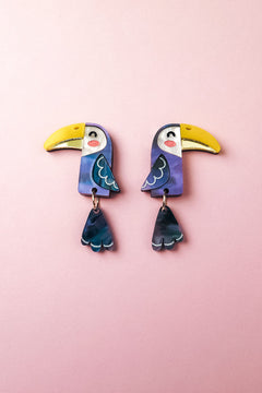 Add a playful pop of color to your look with these handcrafted Toucan Earrings, featuring yellow beaks, blue wings, and dangling blue tail feathers, displayed on a soft pink background.