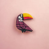 A colorful paper origami toucan pin with a yellow and orange beak and a purple body, handcrafted from eco-friendly materials, is placed on a light pink background.