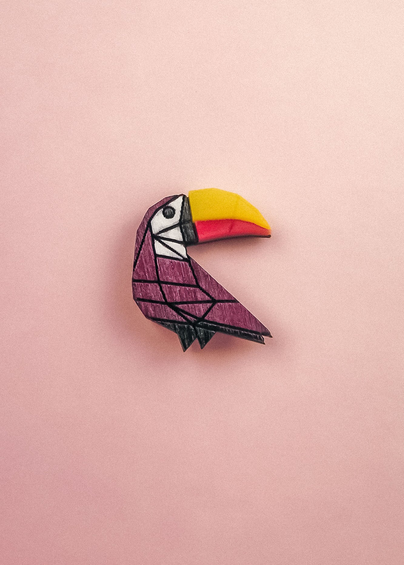 A colorful paper origami toucan pin with a yellow and orange beak and a purple body, handcrafted from eco-friendly materials, is placed on a light pink background.