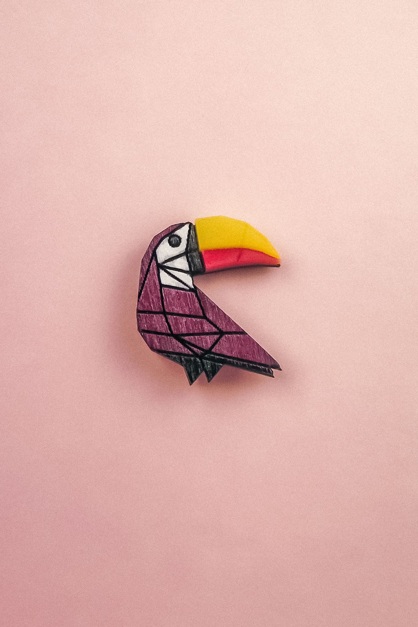 A colorful paper origami toucan pin with a yellow and orange beak and a purple body, handcrafted from eco-friendly materials, is placed on a light pink background.