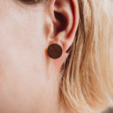 Close-up of a person with short, blonde hair wearing a large, round, handcrafted wooden stud earring in their ear. The focus is on the ear and earring, perfect for elevating any contemporary capsule wardrobe.