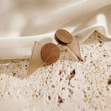 A pair of minimalist earrings with circular wooden tops and translucent triangular bases rest on a textured stone surface beside draped white fabric, perfect for a contemporary capsule wardrobe.