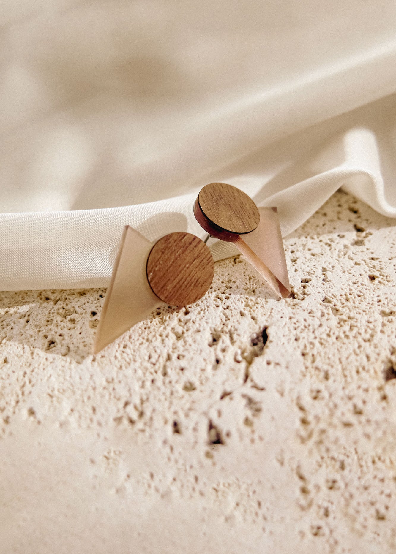 A pair of minimalist earrings with circular wooden tops and translucent triangular bases rest on a textured stone surface beside draped white fabric, perfect for a contemporary capsule wardrobe.
