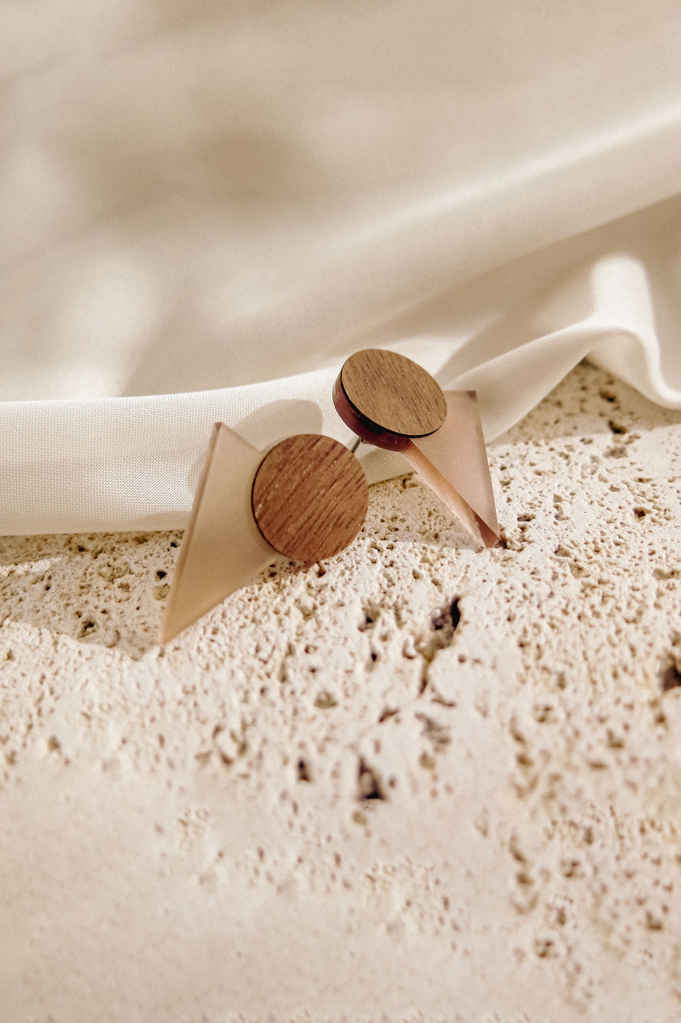 A pair of minimalist earrings with circular wooden tops and translucent triangular bases rest on a textured stone surface beside draped white fabric, perfect for a contemporary capsule wardrobe.