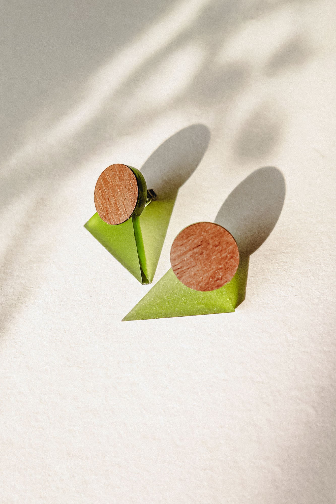 A pair of minimalist triangle earrings with round walnut wood tops and green acrylic glass bases are displayed on a light textured surface, casting soft shadows.
