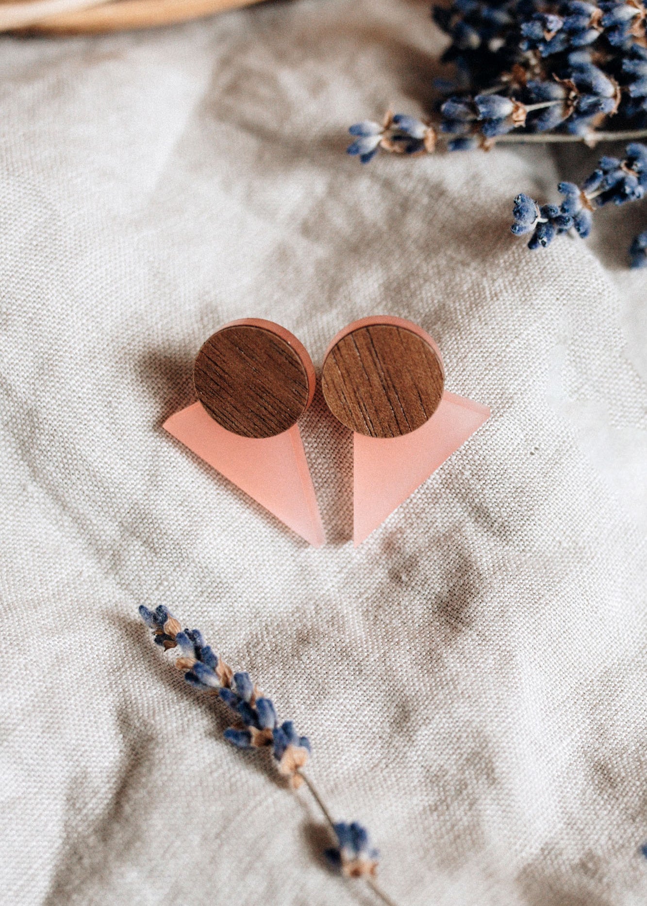 A pair of handcrafted triangle earrings featuring round wooden tops and pink triangular bases are displayed on beige linen fabric, with dried lavender sprigs nearby—perfect for a contemporary capsule wardrobe.