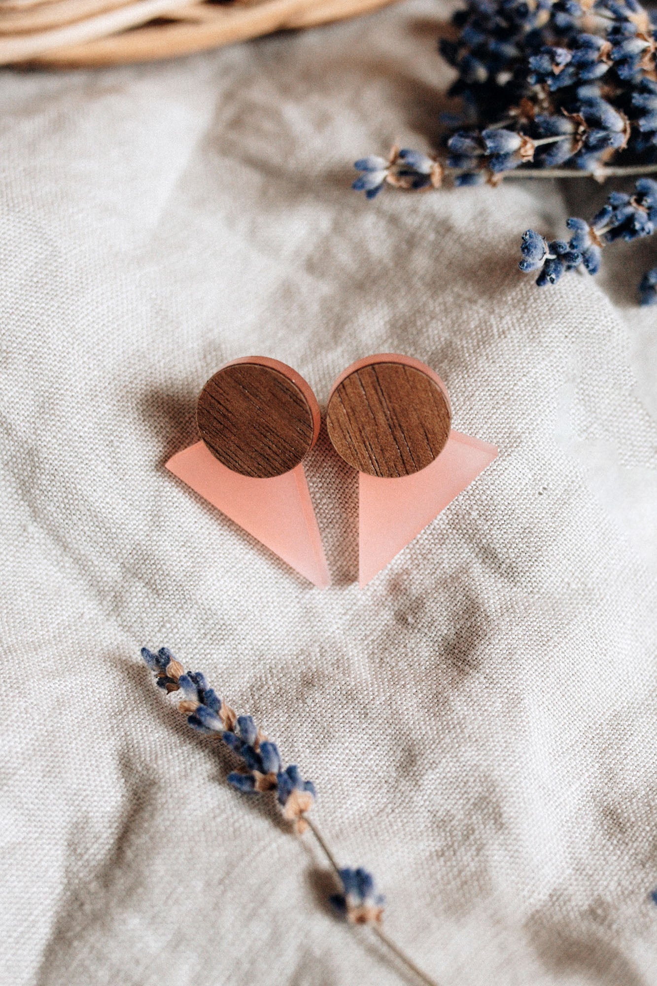 A pair of handcrafted triangle earrings featuring round wooden tops and pink triangular bases are displayed on beige linen fabric, with dried lavender sprigs nearby—perfect for a contemporary capsule wardrobe.