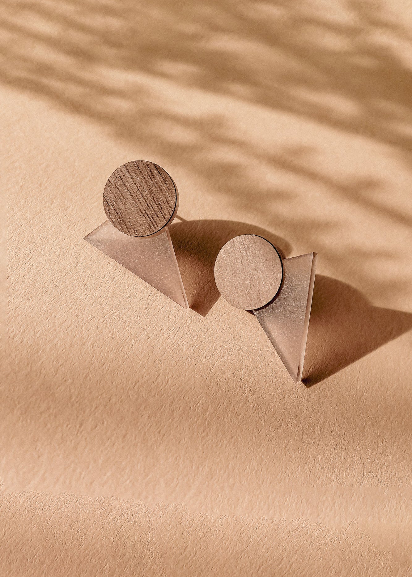 A pair of modern, handcrafted earrings featuring wooden circular tops and translucent triangular bases, displayed on a textured beige surface with soft shadows—perfect for a contemporary capsule wardrobe.