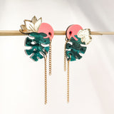The handcrafted Tropicana Earrings feature green monstera leaves, pink circular accents, gold floral details, and dangling gold chains, all brought together in a vibrant tropical-inspired design. Displayed on a horizontal rod against a light background.