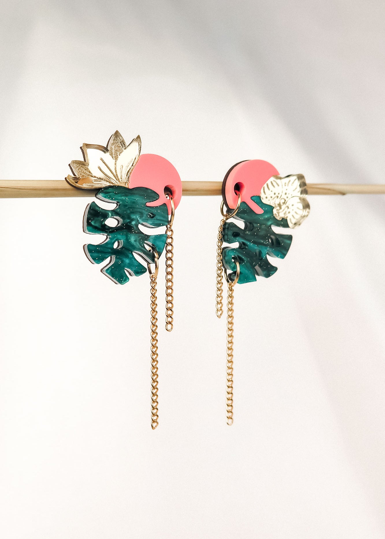 The handcrafted Tropicana Earrings feature green monstera leaves, pink circular accents, gold floral details, and dangling gold chains, all brought together in a vibrant tropical-inspired design. Displayed on a horizontal rod against a light background.