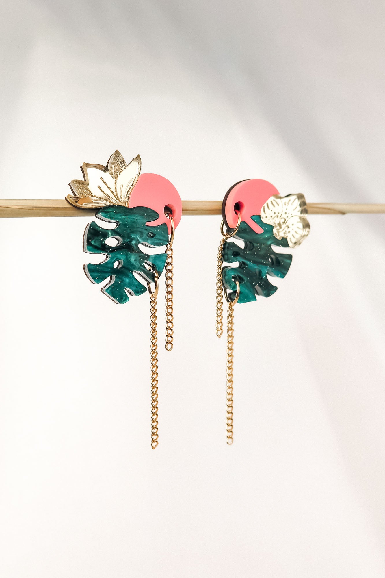 The handcrafted Tropicana Earrings feature green monstera leaves, pink circular accents, gold floral details, and dangling gold chains, all brought together in a vibrant tropical-inspired design. Displayed on a horizontal rod against a light background.